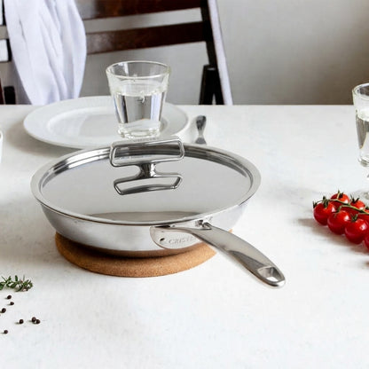 The Cristel Castel'Pro 9.5" deep frying pan with its custom-fit stainless steel lid, resting on a white table setting with fresh tomatoes and herbs.
