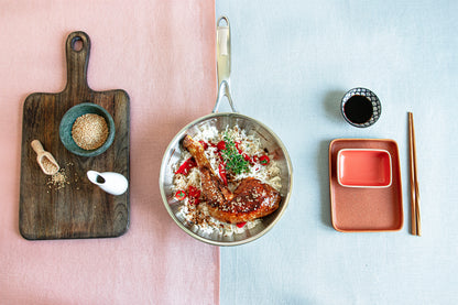 Top-down editorial view of the Cristel Castel'Pro deep frying pan filled with glazed chicken and rice, styled with a wooden cutting board and chopsticks.